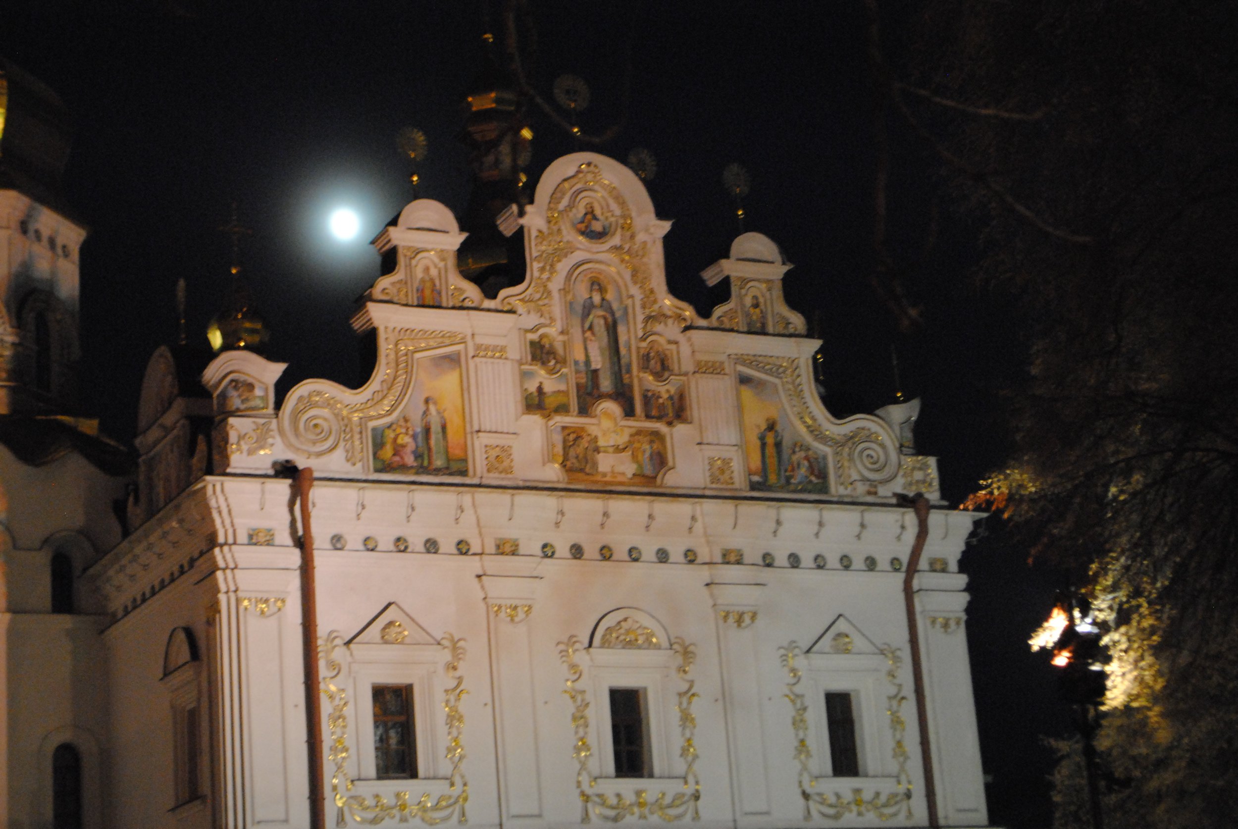 Nighttime view of an ornate white building with gold accents, decorated with religious paintings and topped with spires, illuminated by external lighting. The moon is visible in the dark sky.