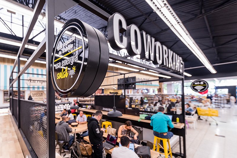 People working at tables and on laptops inside a cafe or co-working space within an indoor marketplace or shopping mall. Large sign overhead reading 'COWORKING' with a clock showing 'Energía sale' in Spanish.