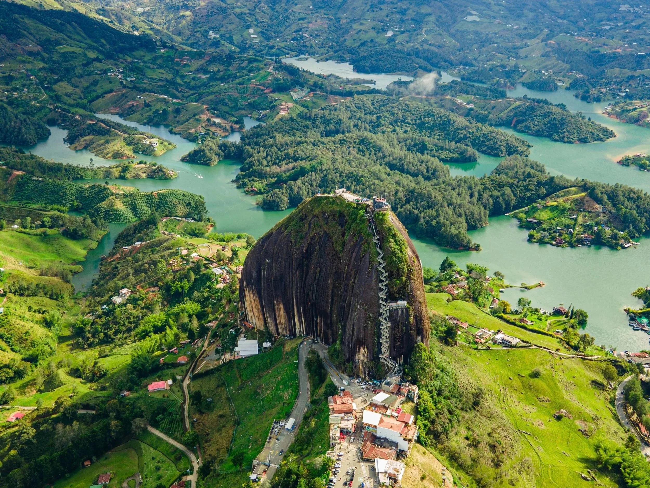 Aerial view of a lush green landscape featuring a large mountain with a staircase zigzagging up its side, surrounded by winding rivers, trees, and scattered houses.
