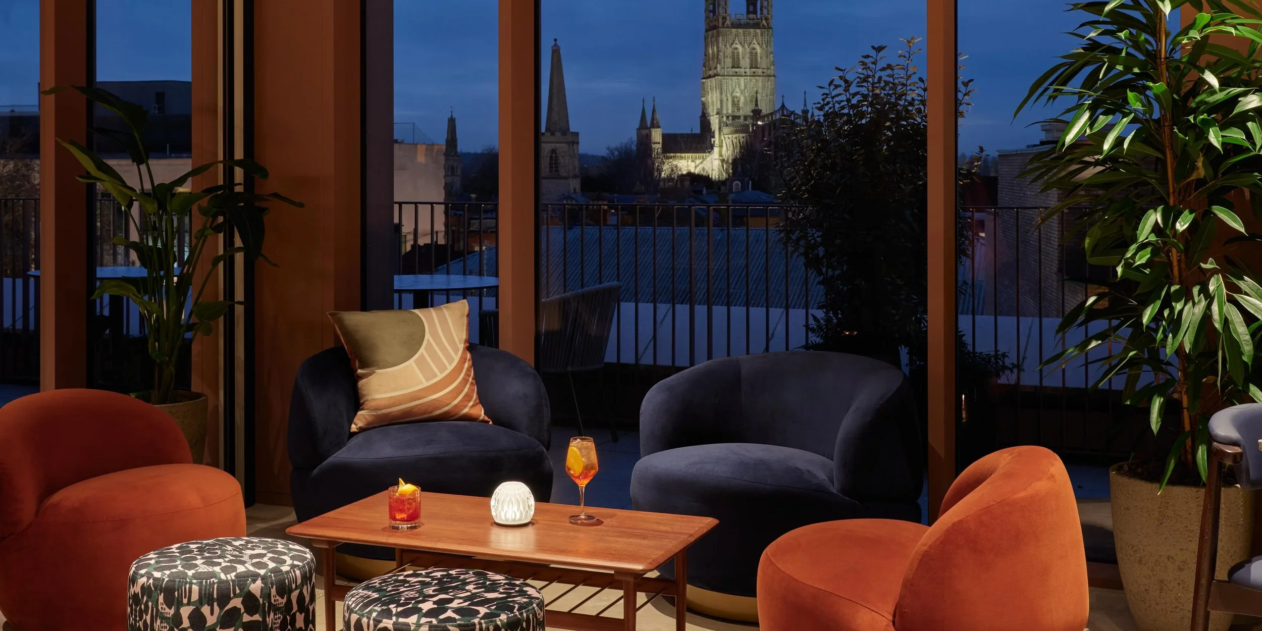 Interior of a living room with large windows showing a cityscape at dusk, including illuminated church steeples and towers, decorated with plants and modern furniture.