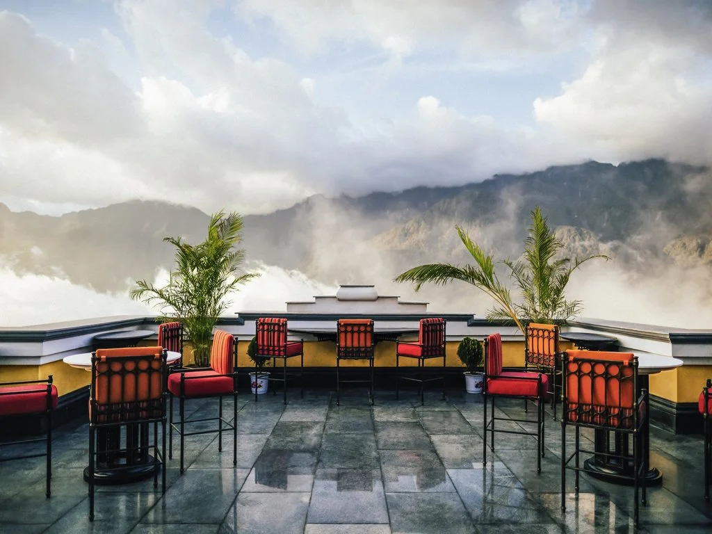 A rooftop terrace with red chairs and potted plants overlooking mountains and clouds.