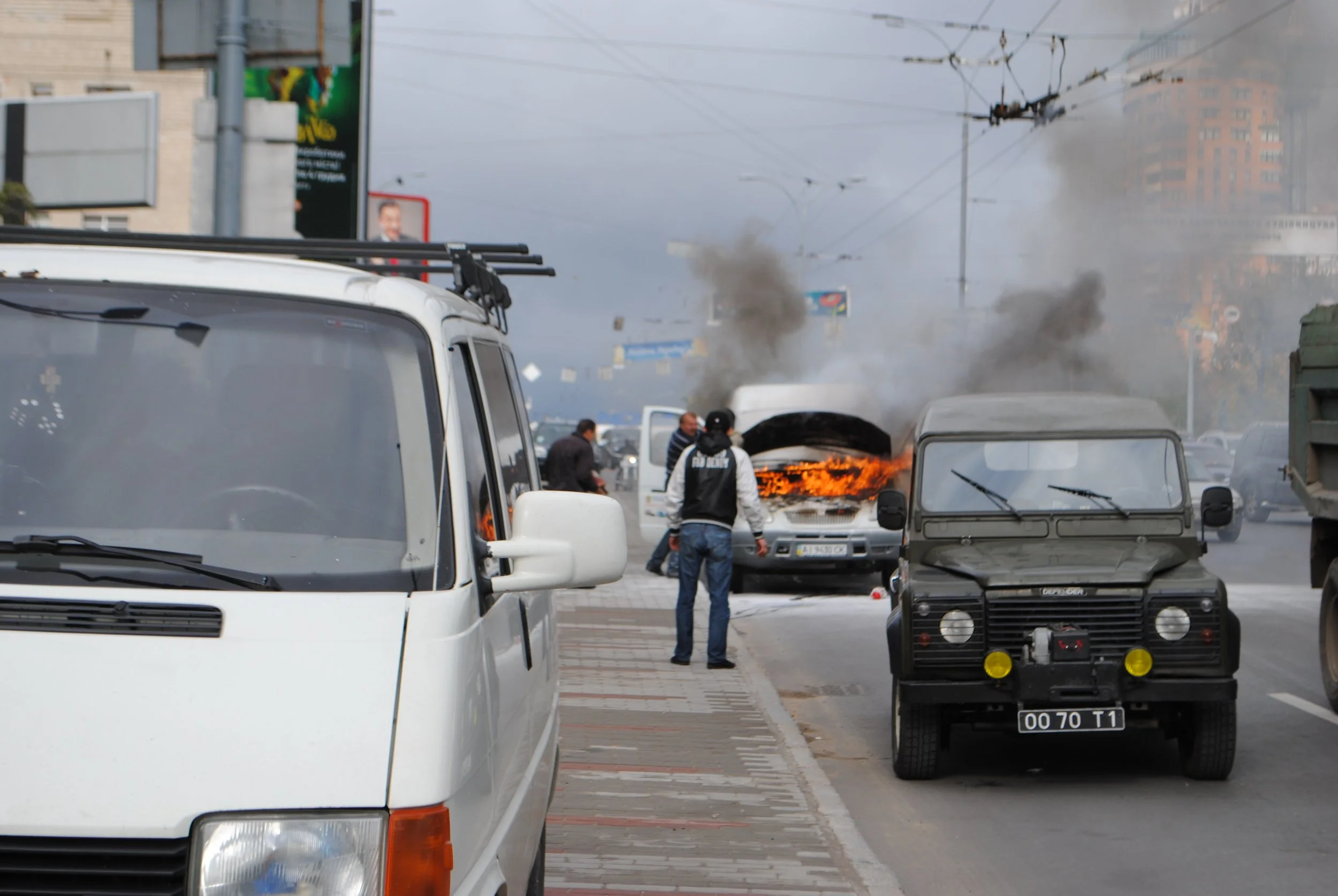A street scene with a vehicle on fire, thick black smoke rising, and several people nearby, in an urban area with multiple vehicles and buildings in the background. Kiev November 2011, Marina Parrish ©