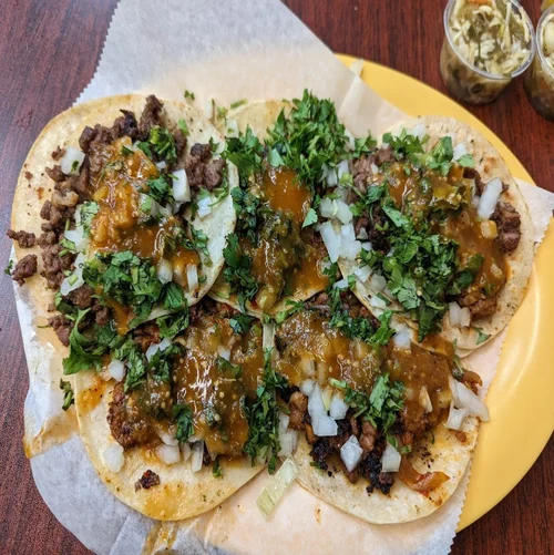 Five beef tacos topped with chopped onions and cilantro, with green and red sauce drizzled on top, served on a yellow plate with two cups of shredded lettuce in the background.