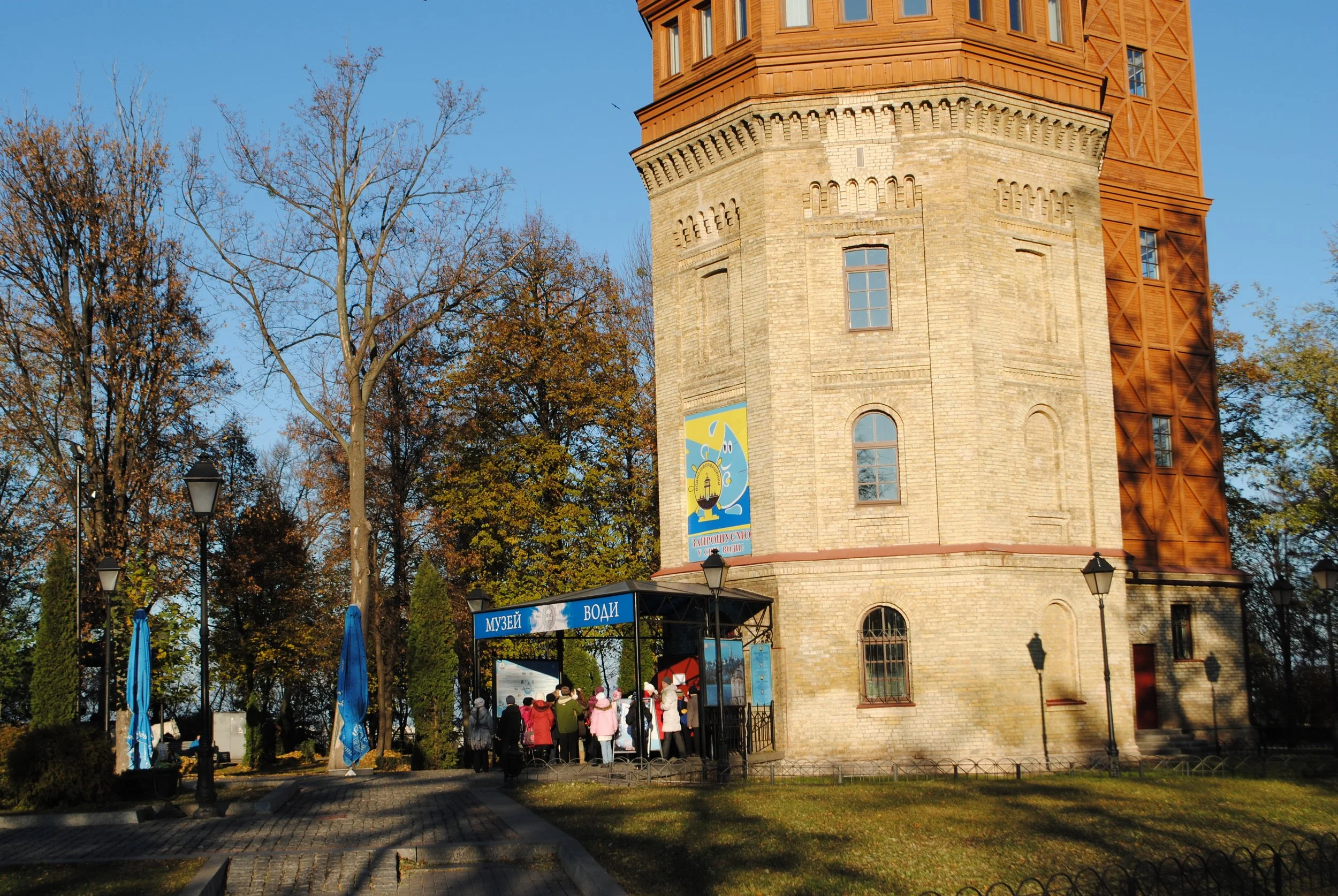 A historic brick water tower with a wooden observation deck on top, surrounded by autumn trees and a group of people gathered near the entrance under signs that read 'МУЗЕЙ ВОДИ'.