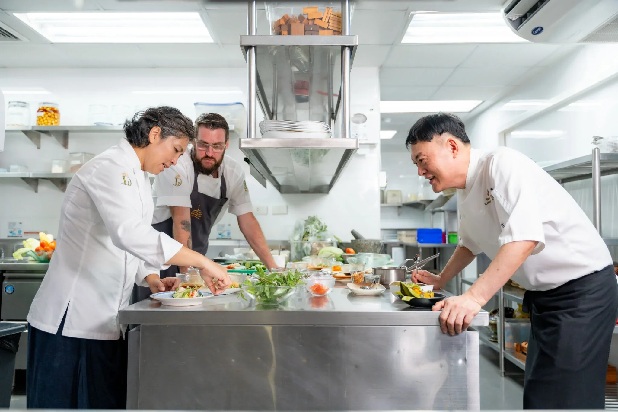 Three chefs in white uniforms preparing dishes in a professional kitchen with stainless steel countertops and shelves filled with ingredients.