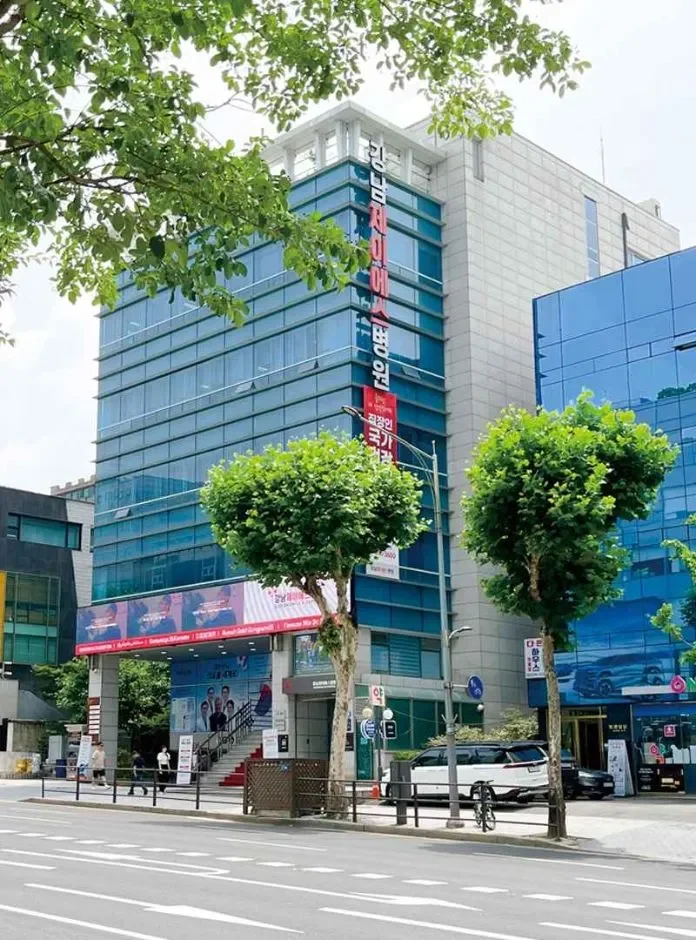 Modern multi-story office building with Korean signage, glass exterior, and adjacent similar structures on a city street with trees and parked cars.