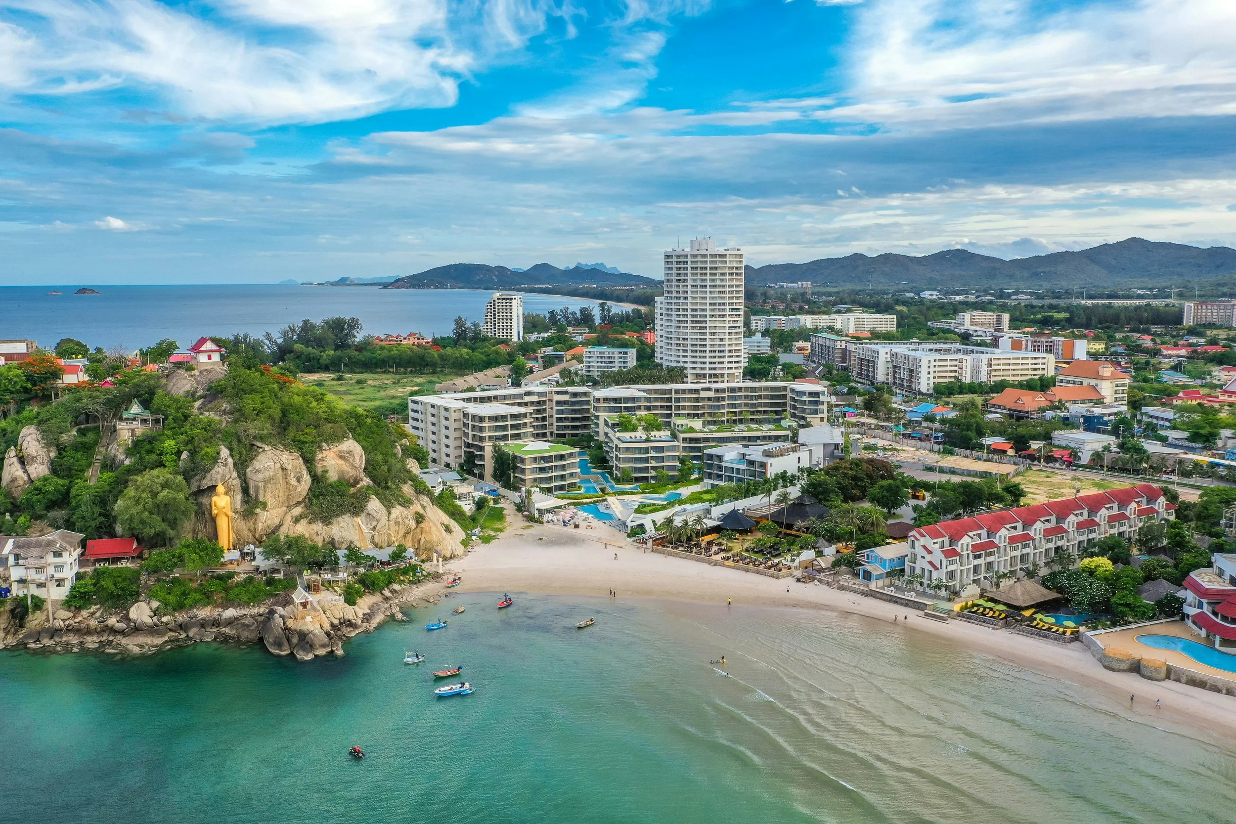A scenic aerial view of a coastal city featuring high-rise buildings, a small beach, lush greenery, and islands in the distance under a partly cloudy sky.