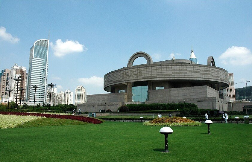 Modern cityscape with a unique circular building in the foreground, surrounded by green lawns, flowerbeds, and high-rise skyscrapers under a blue sky.