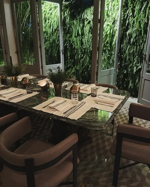 A dining table set with white napkins, glasses of water, and small flower arrangements, in a room with large windows showing lush green foliage outside.