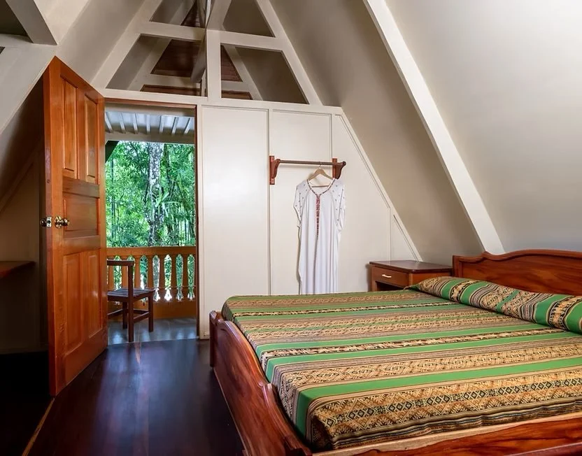 Bedroom with wooden furniture, a bed with a patterned bedspread, a small wooden dresser, a white dress hanging on a wall hook, and an open door leading to a balcony with trees outside.