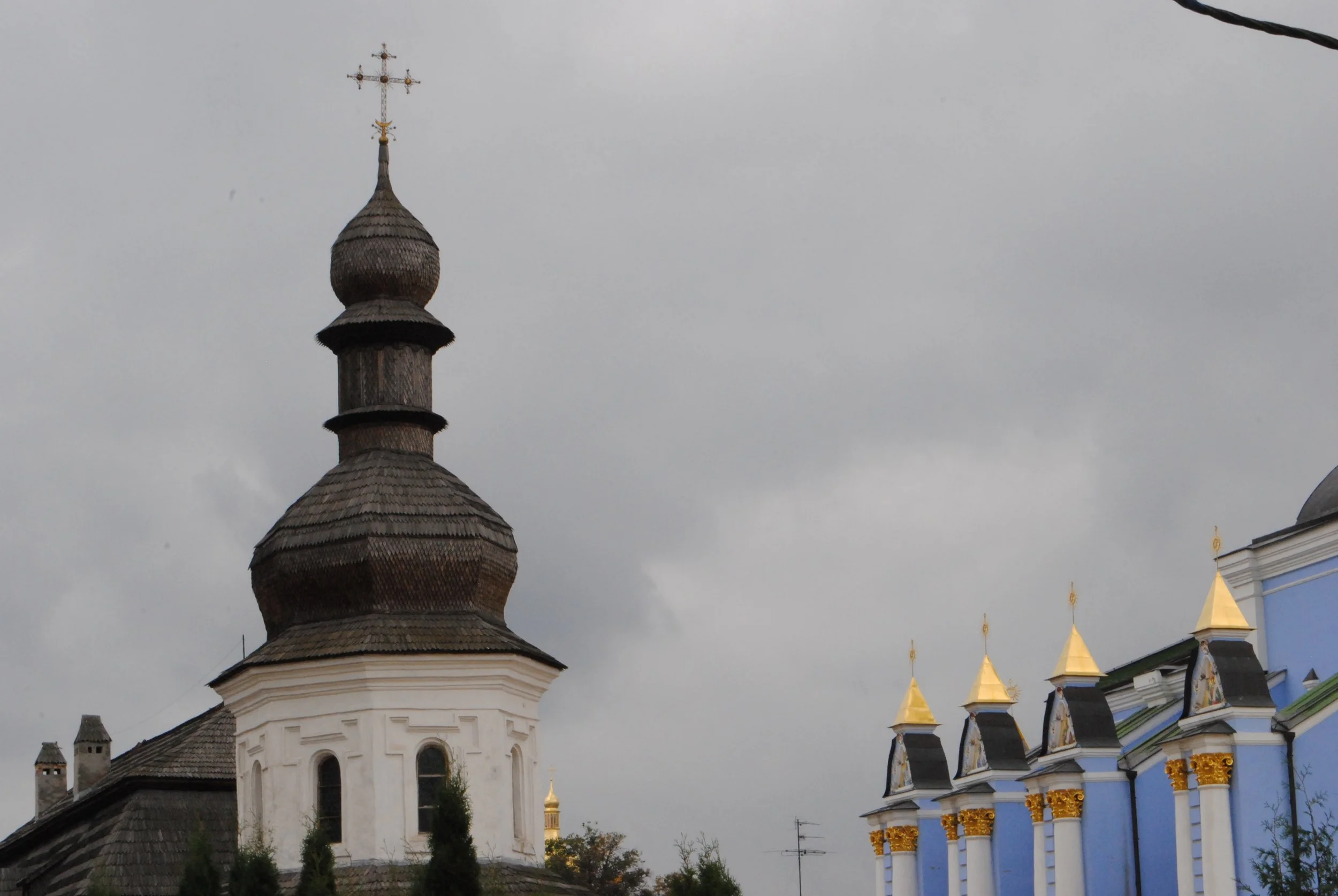 A church tower with a wooden shingle roof and a golden cross on top, alongside a building with golden domes and blue and white walls, located under an overcast sky.