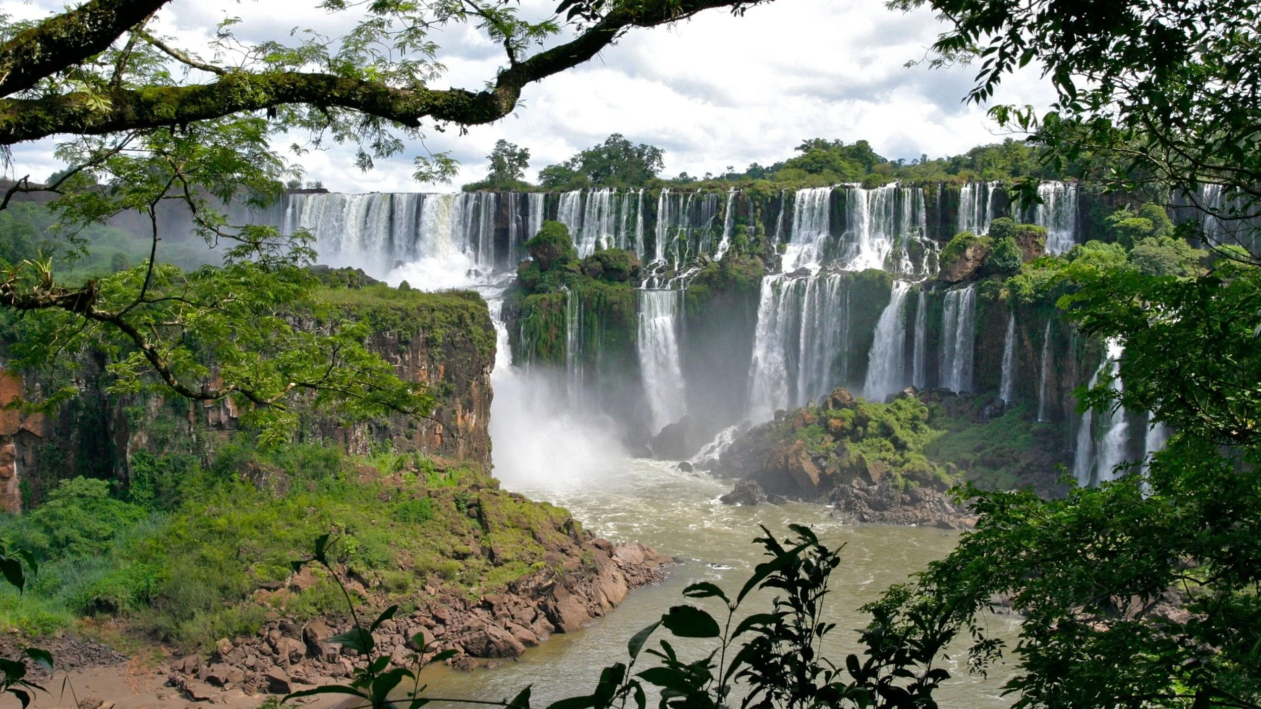 A large waterfall flowing over a cliff surrounded by lush green trees and foliage, with mist rising at the base.
