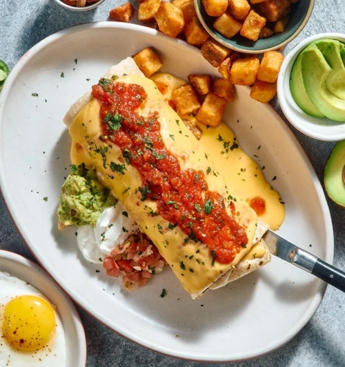 Breakfast dish with stuffed burrito topped with cheese, red sauce, and chopped herbs, served with guacamole, sour cream, home fries, and avocado slices.