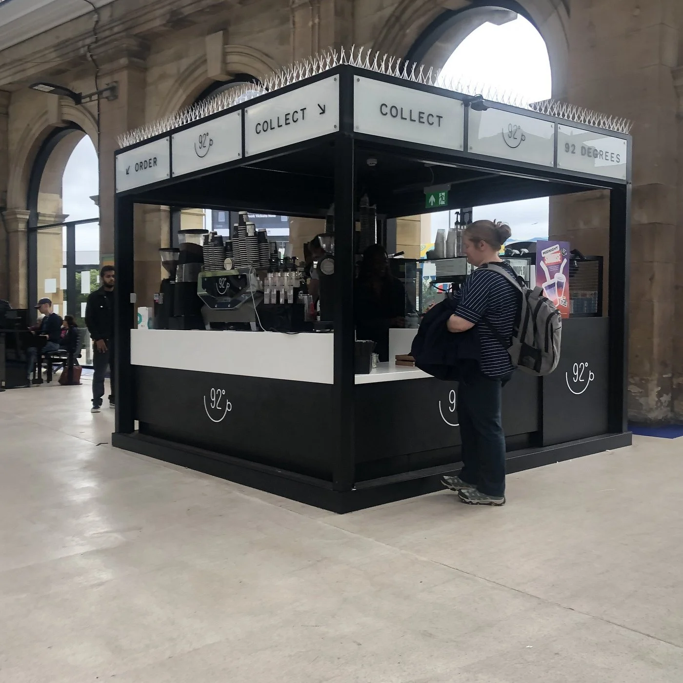 A small coffee kiosk inside a train station, serving coffee and beverages. A woman with a backpack is waiting at the counter, and a barista is behind the counter preparing drinks. The kiosk has signs indicating to order and collect, with the temperat