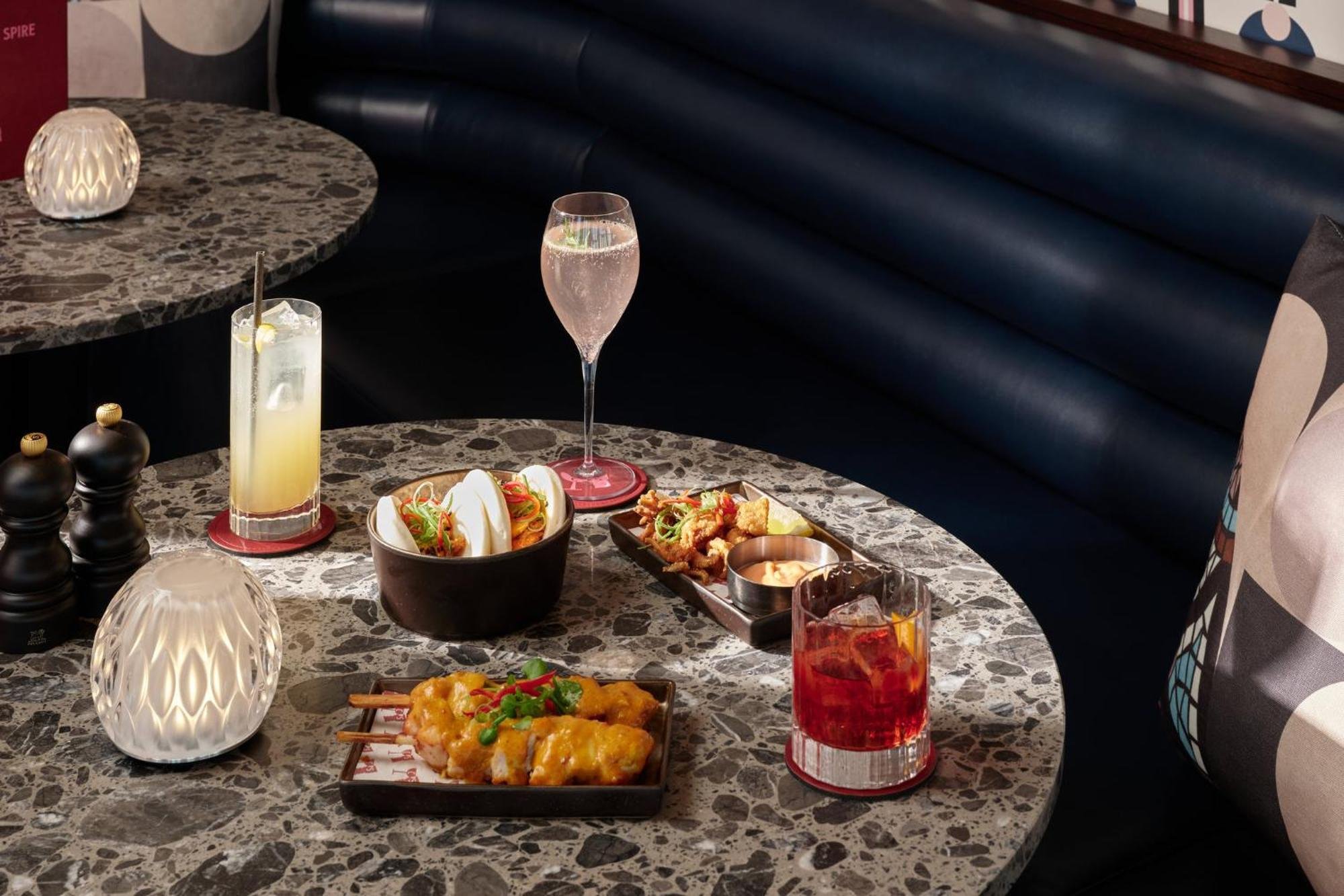 Table with various dishes and drinks, including sushi, fried food, and beverages in a restaurant setting, with a black leather banquette and decorative lamps.