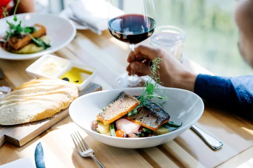 A dining table with a plate of grilled fish, vegetables, and garnishes, a glass of red wine, slices of bread with butter, and condiments on the side.