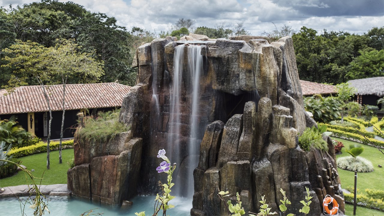 A large artificial waterfall flowing over a rock formation into a pool, surrounded by lush greenery and garden structures.