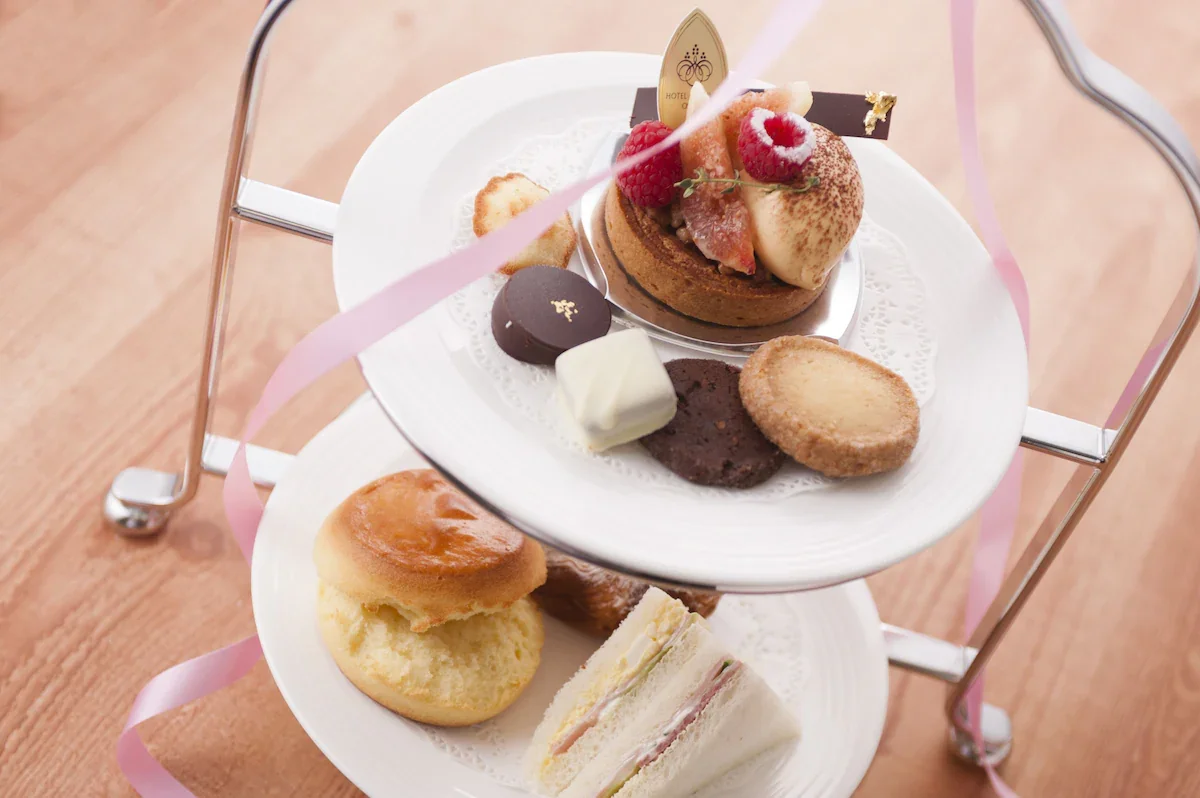 Two-tiered tray with assorted desserts, including fruit-topped cake, cookies, chocolates, a cupcake, and a sandwich, on a wooden table with pink ribbons.