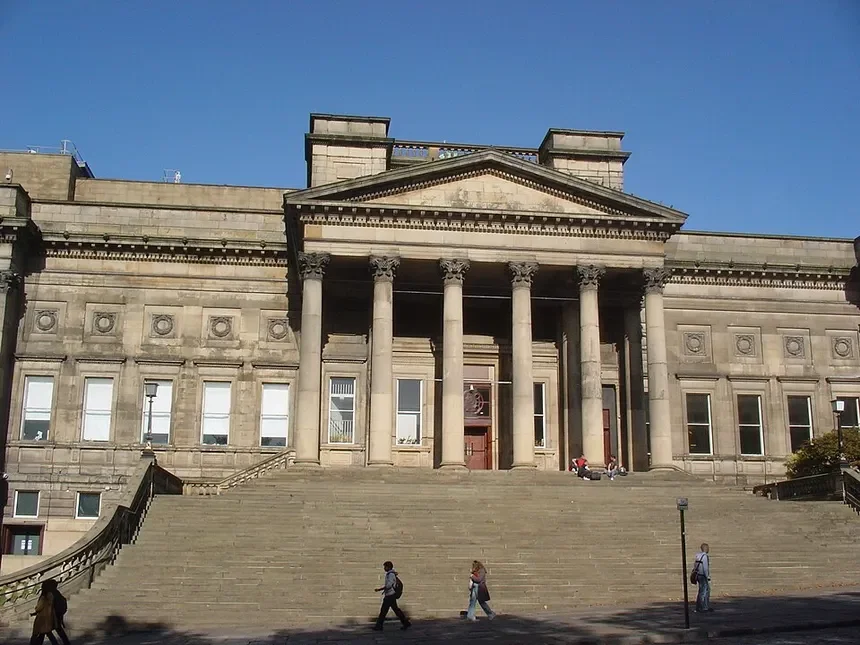 A large historic building with classical architecture, featuring tall columns, a triangular pediment, and stone steps leading up to the entrance, under a clear blue sky.
