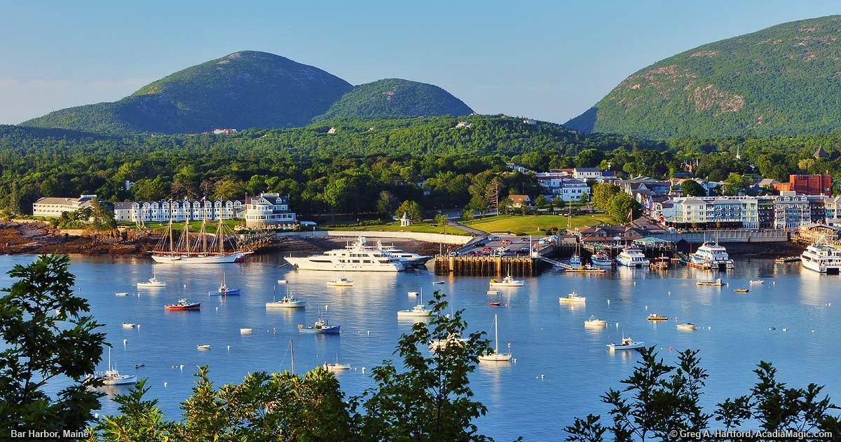 Scenic view of Bar Harbor, Maine, featuring a harbor with numerous sailboats and yachts, lush green trees in the foreground, a row of buildings along the shoreline, and green mountains in the background under a clear blue sky.