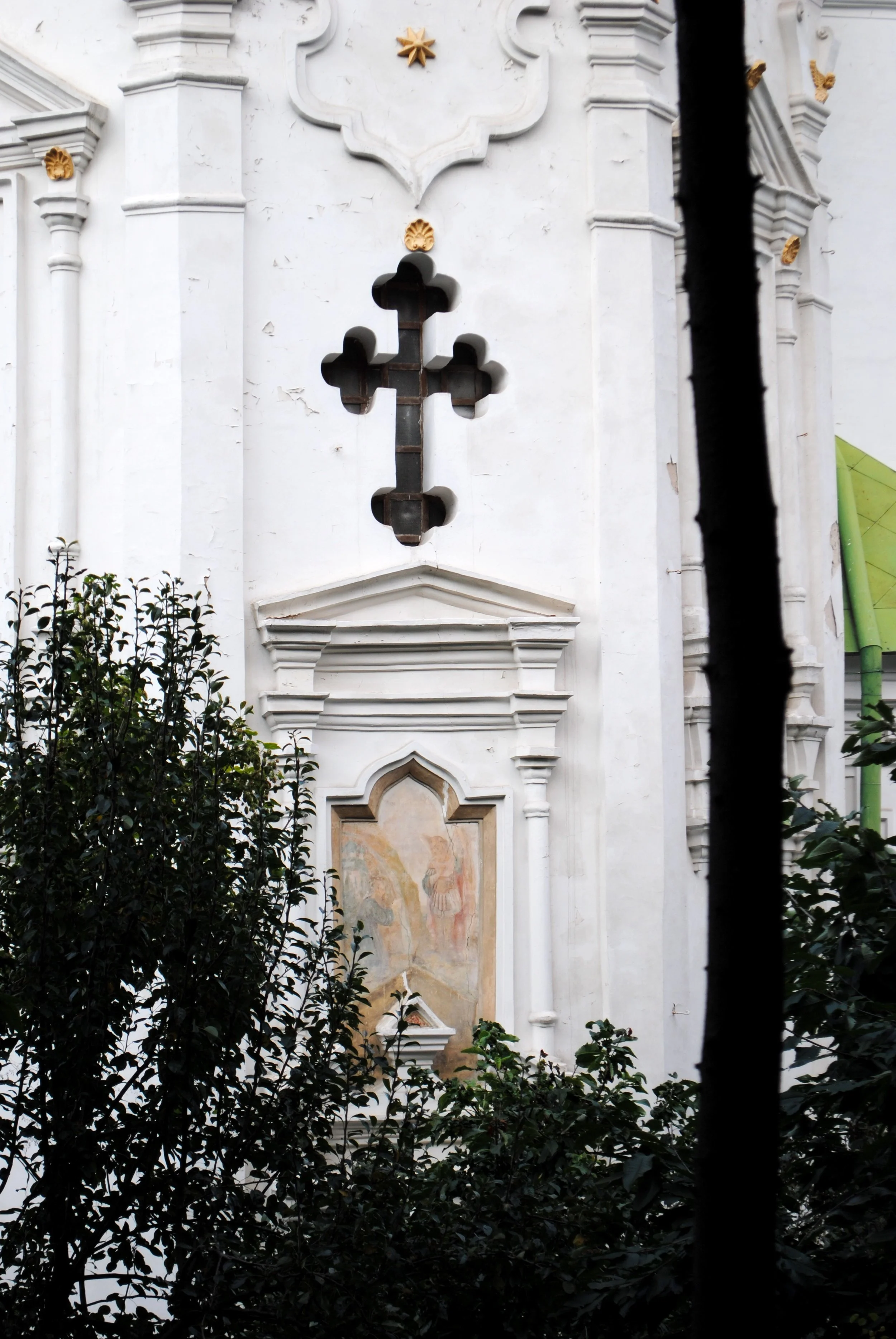 A white building with architectural details and a decorative cross-shaped window, partially obscured by leaves and branches in the foreground.