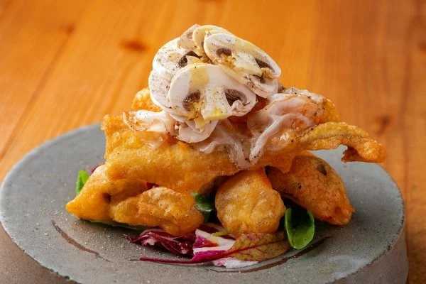 Fried fish fillet topped with chopped onions and drizzled with sauce, served on a gray plate with leafy greens, on a wooden table.