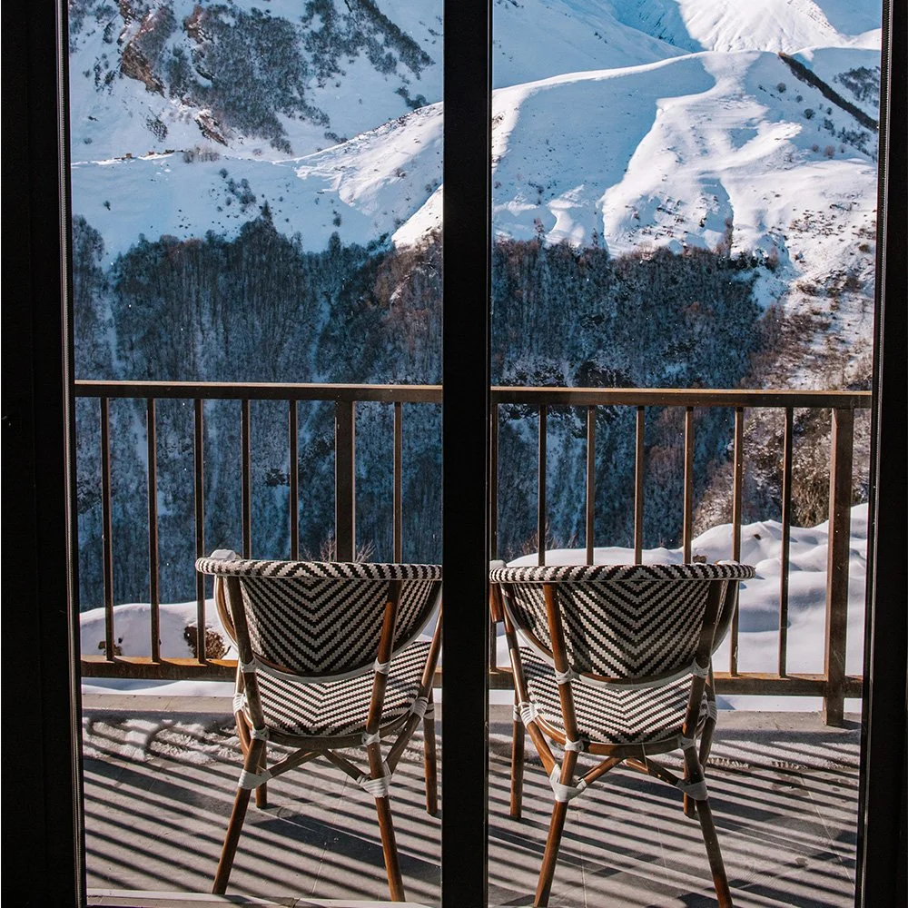 Snow-covered mountain landscape seen through a glass window with two chairs on a balcony.