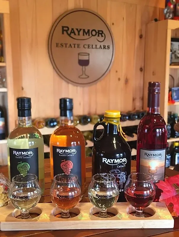 Four bottles of Raymor estate cellar wines and four tasting glasses with different colored wines arranged on a wooden tasting tray.