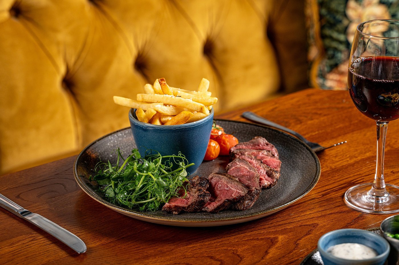 A plate with sliced steak, fresh greens, roasted cherry tomatoes, and a blue cup of French fries. There is a glass of red wine on a wooden table with a black fork and a small bowl of white sauce or dip.