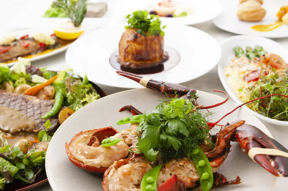 Assorted gourmet seafood dishes and salads on white plates and bowls, featuring lobster, salads, and various appetizers, with a focus on a lobster dish garnished with herbs and vegetables.