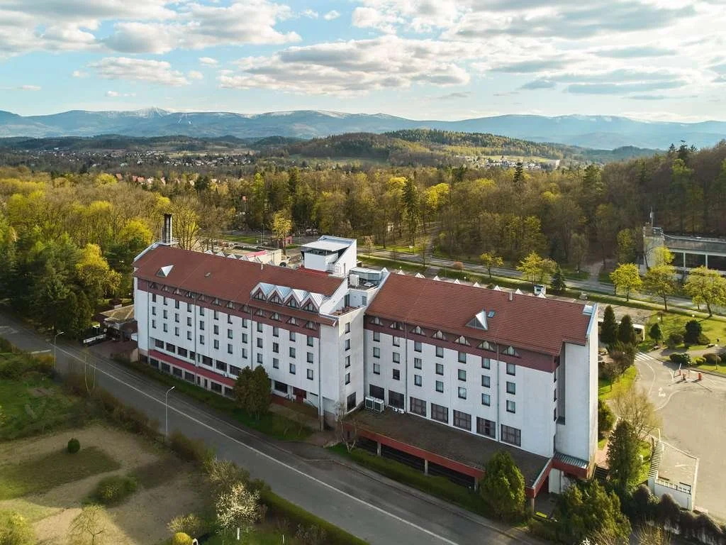 Aerial view of a multi-story white hotel with a red roof, surrounded by trees, roads, and mountains in the background on a partly cloudy day.