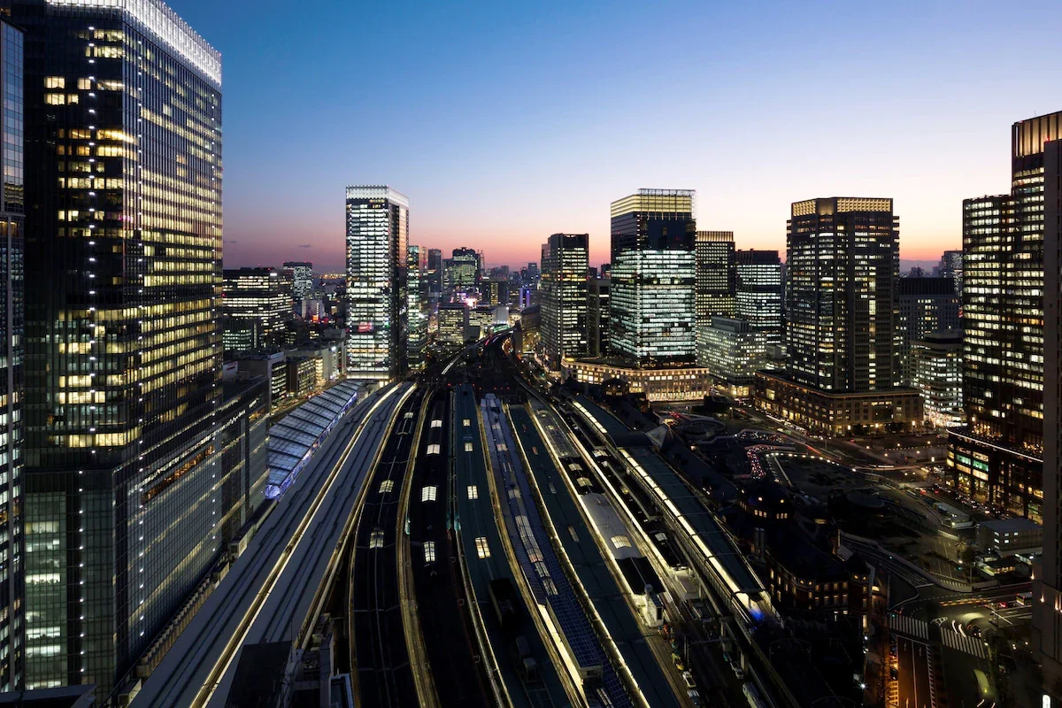 Cityscape of a modern downtown area at dusk with illuminated skyscrapers, train tracks in the foreground, and a clear sky transitioning from sunset to night.
