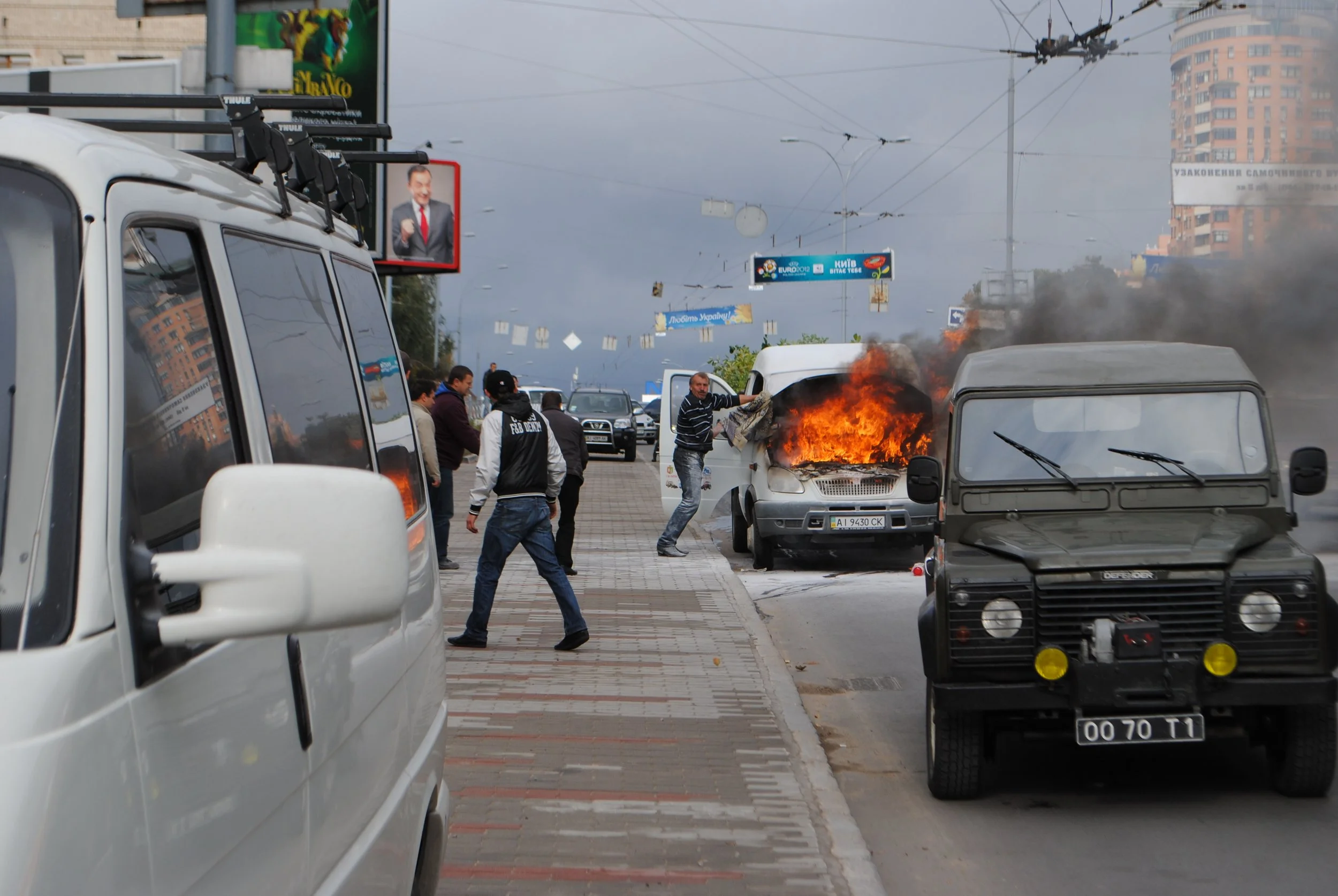 A car on fire with flames and smoke coming from the hood on a city street, while several pedestrians observe the scene. Vehicles and buildings are visible, with a man actively pouring a liquid on the fire. Kiev November 2011, Marina Parrish ©