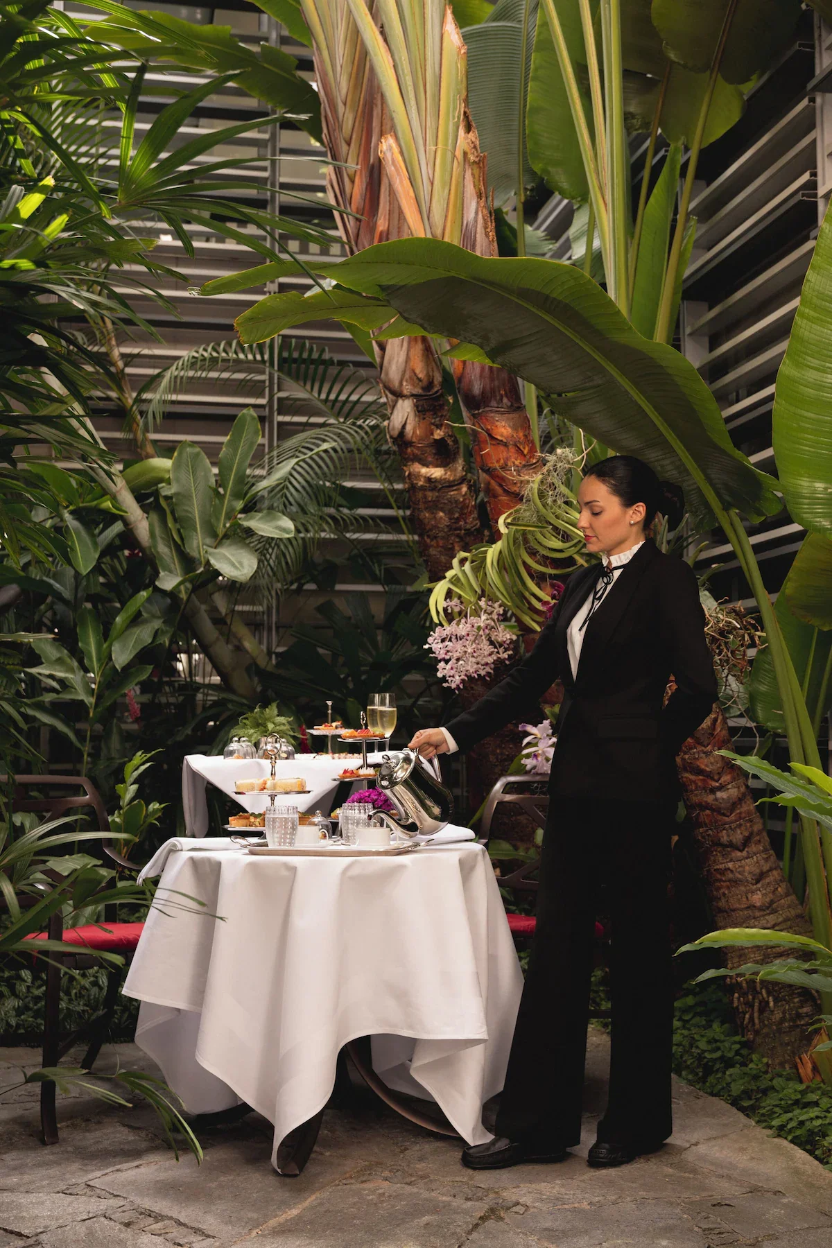 A waitress in a black suit pouring tea at a table set for tea with a tiered tray of desserts, surrounded by lush green tropical plants in a greenhouse or conservatory.
