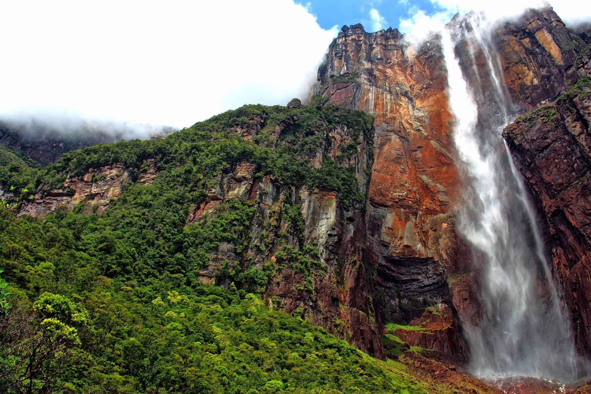 A tall waterfall cascading down a rocky cliff in a lush green mountainous landscape, with mist and clouds around the top of the cliff.