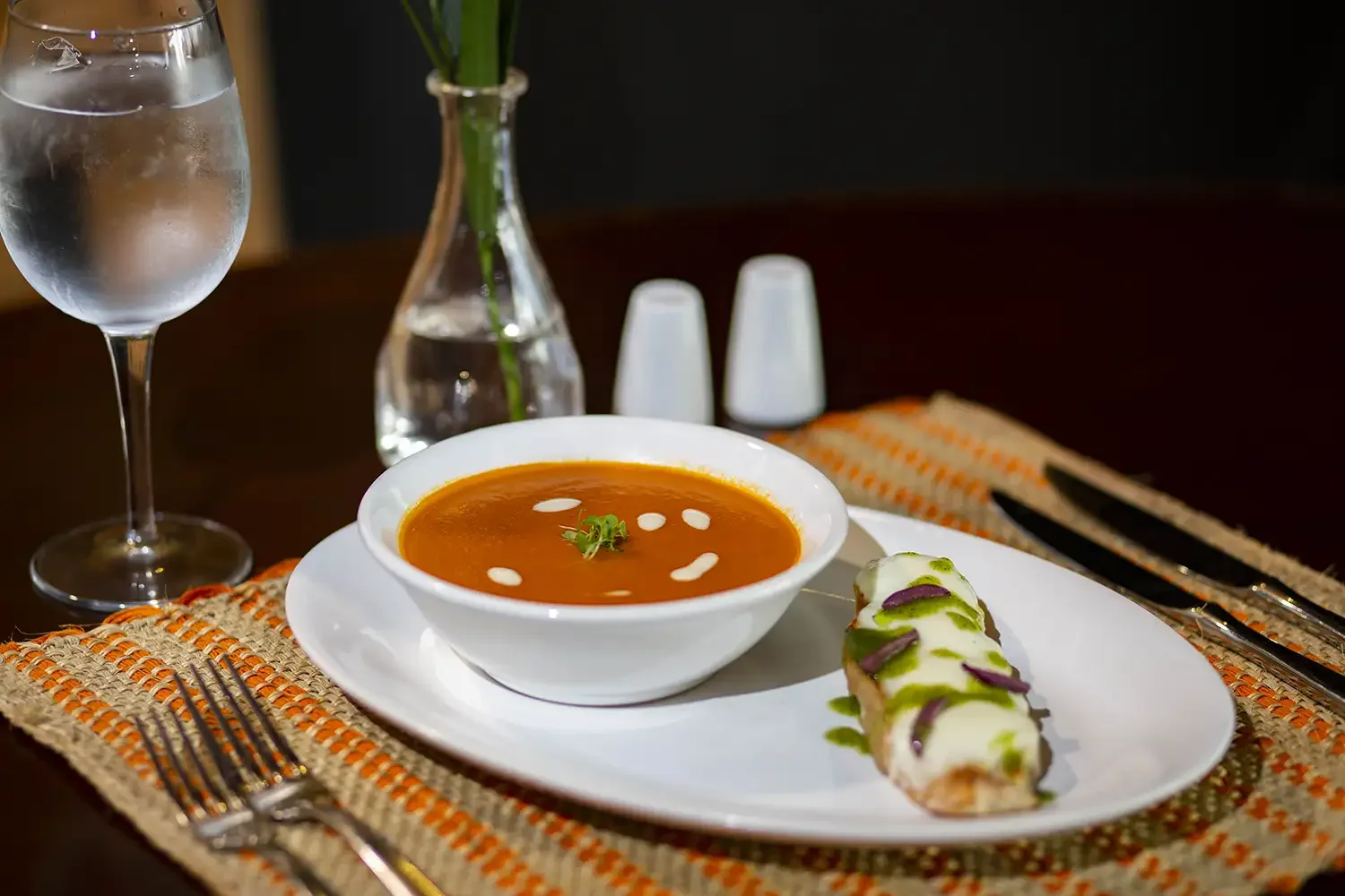A bowl of tomato soup garnished with herbs and drizzled with cream, served with an appetizer of avocado toast topped with herbs, on a white plate with cutlery, a glass of water, a small vase with green plant, and salt and pepper shakers on a woven or