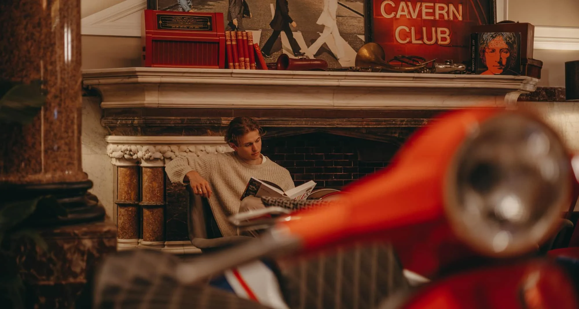 Young person sitting on the floor reading a book beneath a fireplace mantel decorated with vintage items, including a horn, book, and a painting of The Beatles, with a 'CAVERN CLUB' sign illuminated in the background.