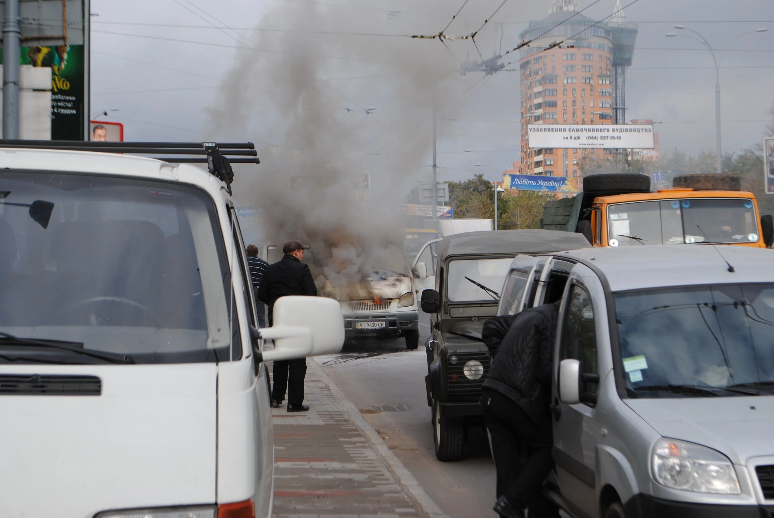 A vehicle caught on fire with black smoke rising in an urban street, surrounded by parked cars and a few people nearby, with a tall building in the background. Kiev November 2011, Marina Parrish ©