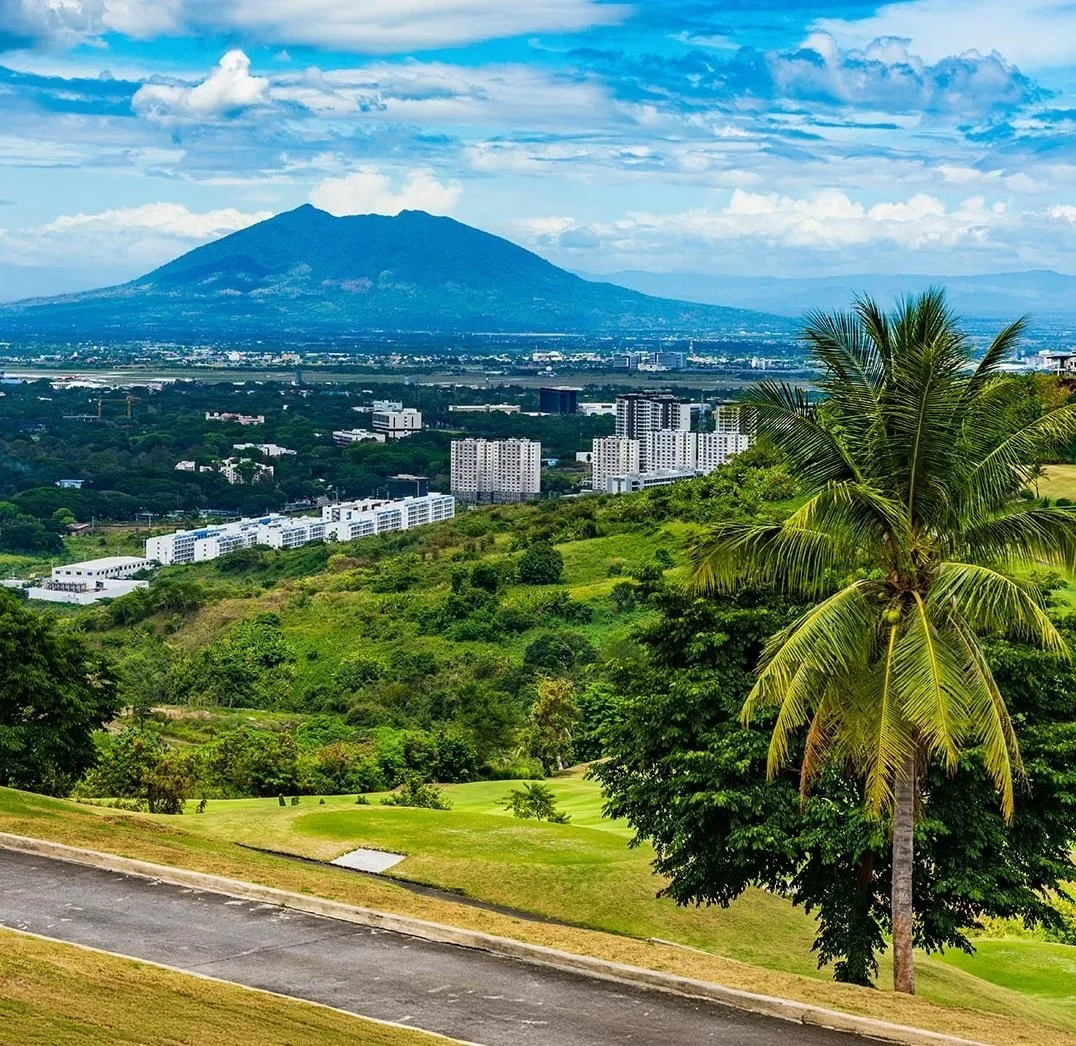 A scenic view of a tropical landscape with green hills, palm trees, and urban buildings in the foreground. In the background, there is a large mountain under a partly cloudy sky.