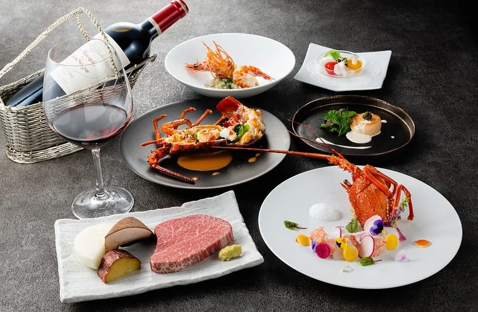 A luxurious spread featuring a glass of red wine with a bottle in a basket, various gourmet dishes including lobster, steak, and assorted appetizers, all presented on elegant plates with colorful garnishes, set on a dark surface.