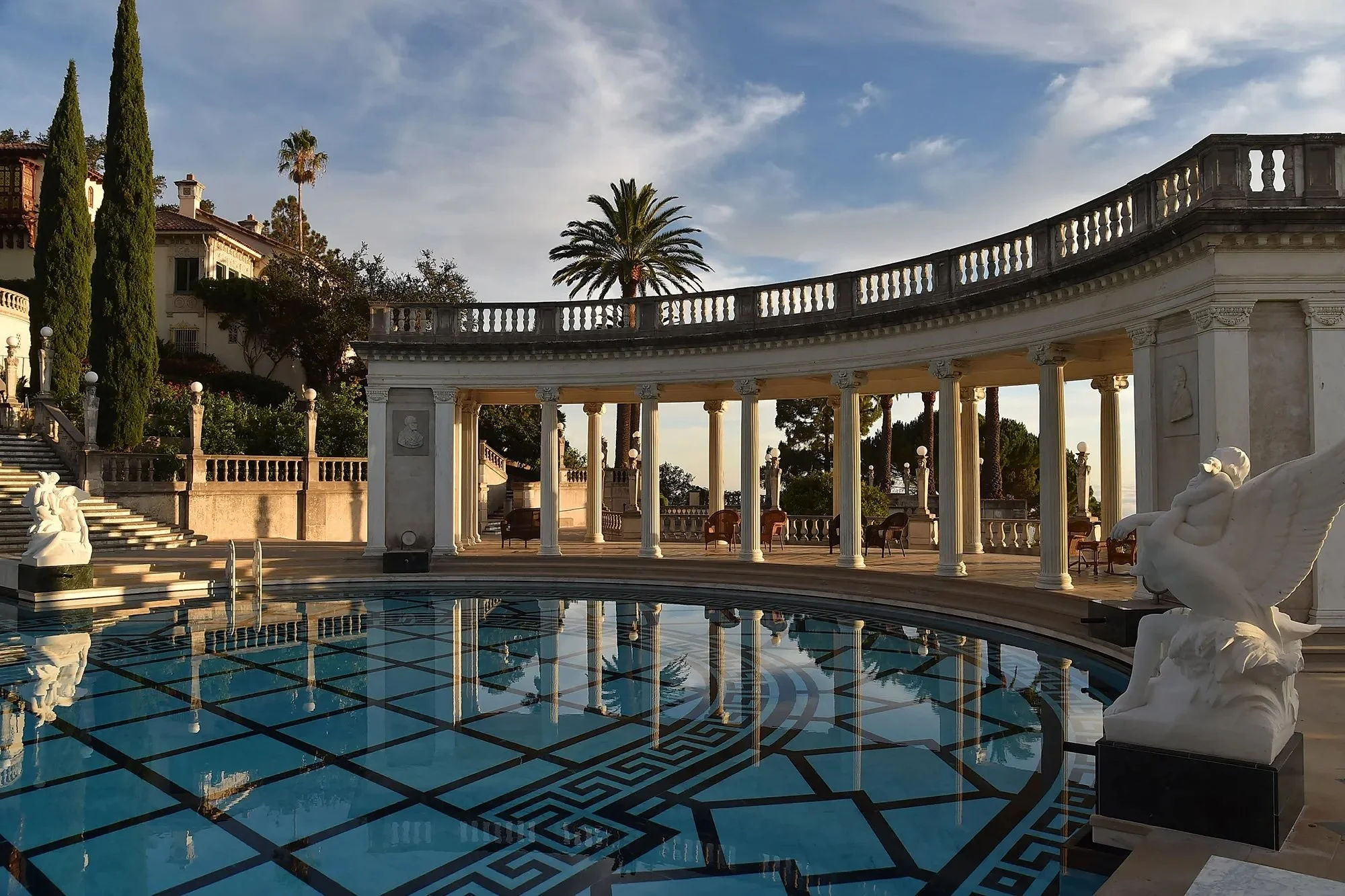 Luxury outdoor pool area with marble statues, a colonnade, palm trees, and elegant furniture under a blue sky.