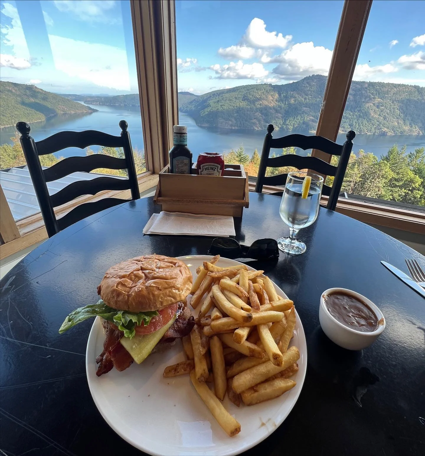 A burger with lettuce, tomato, bacon, and cheese, served with French fries and a side of baked beans on a white plate, on a black table with a scenic lake view through large windows in the background.