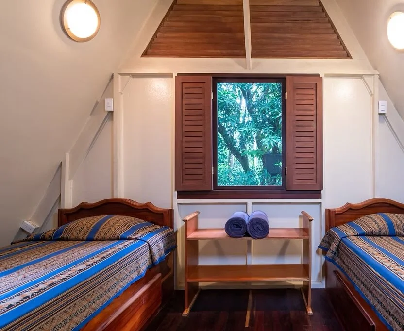 Room with two twin beds with striped bedding, a wooden table with two rolled towels, a window with open wooden shutters showing green foliage outside, and two wall-mounted lights.