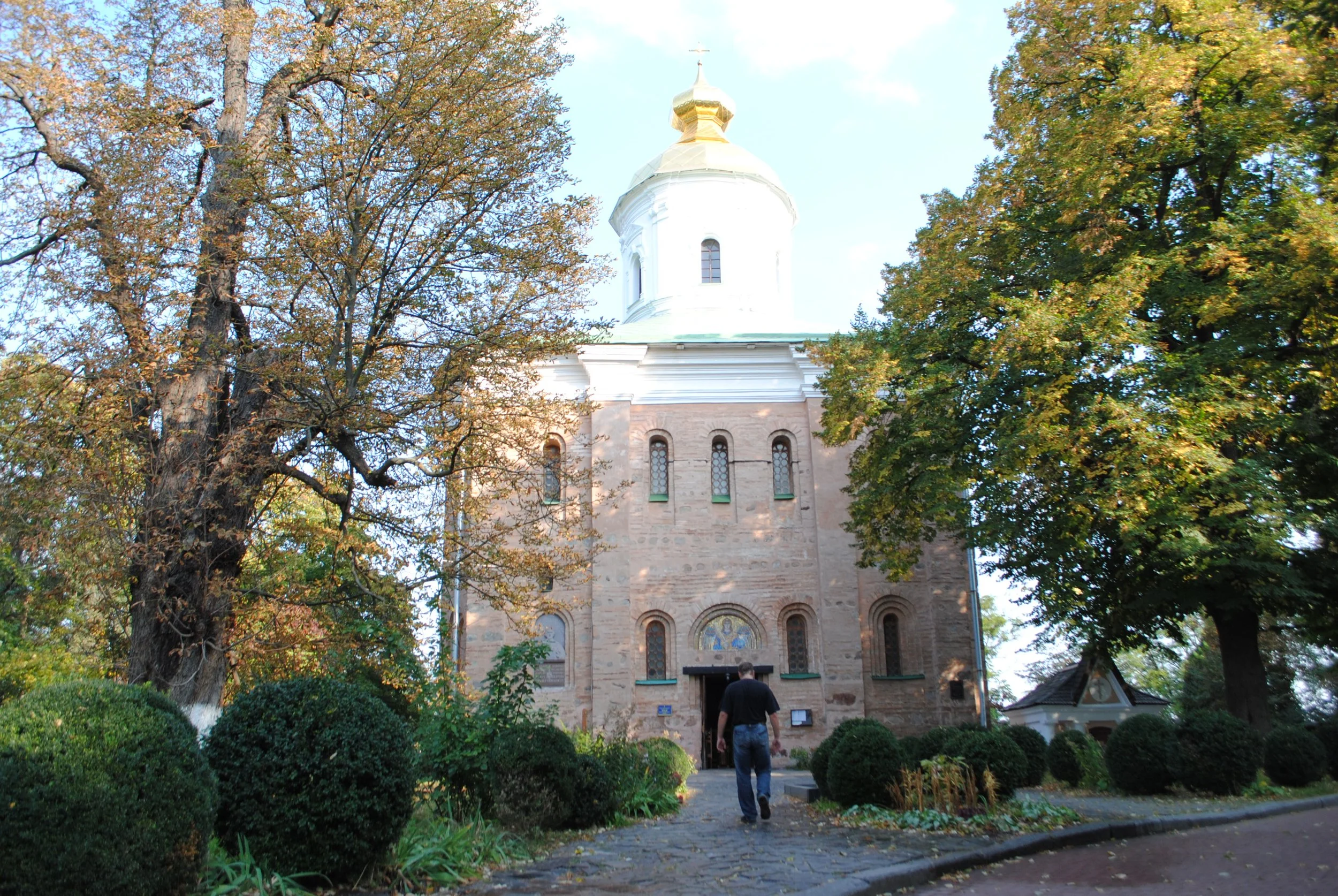 A church surrounded by trees with autumn foliage, a person walking toward it, and well-maintained bushes and plants in the foreground.