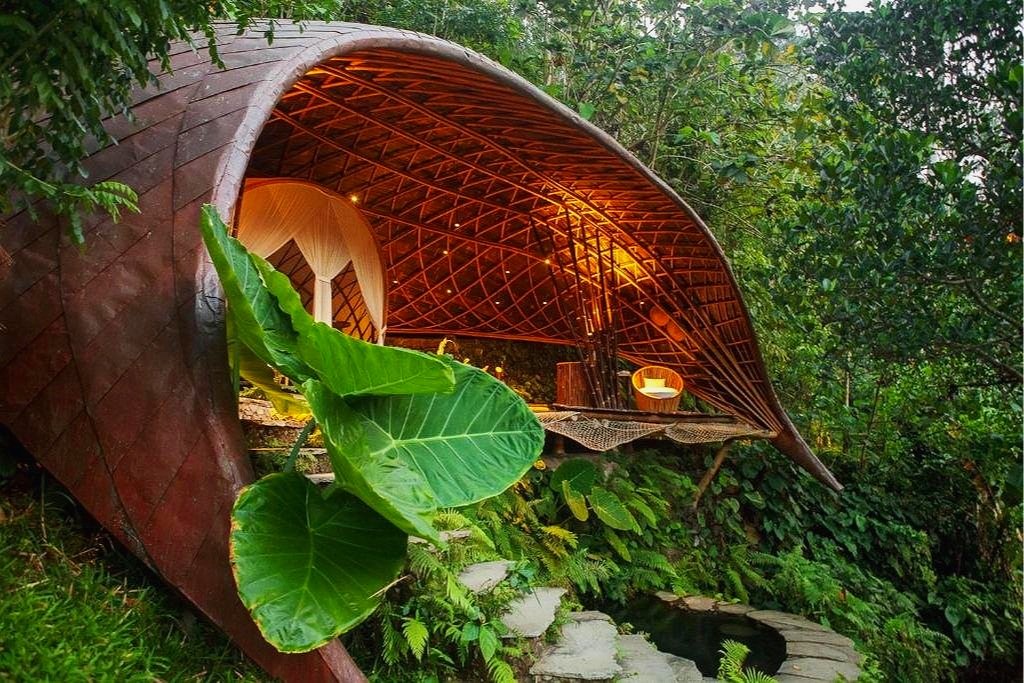 A treehouse with a curved, leaf-like roof and open walls, surrounded by lush green foliage, with cozy seating inside.
