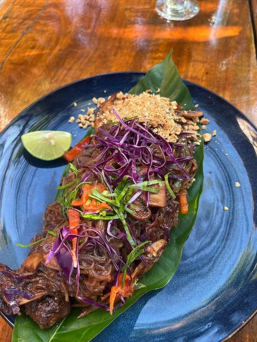 A plate of slow-cooked beef with shredded purple and green cabbage, carrots, and fresh herbs, garnished with crushed peanuts and a lime wedge, served on a banana leaf on a blue plate.