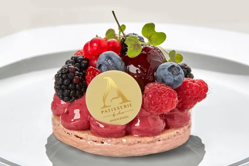 A dessert tart topped with assorted fresh berries, a cherry, and a small green leaf, with a logo that reads 'Patisserie by Anna Baden-Baden' on top.