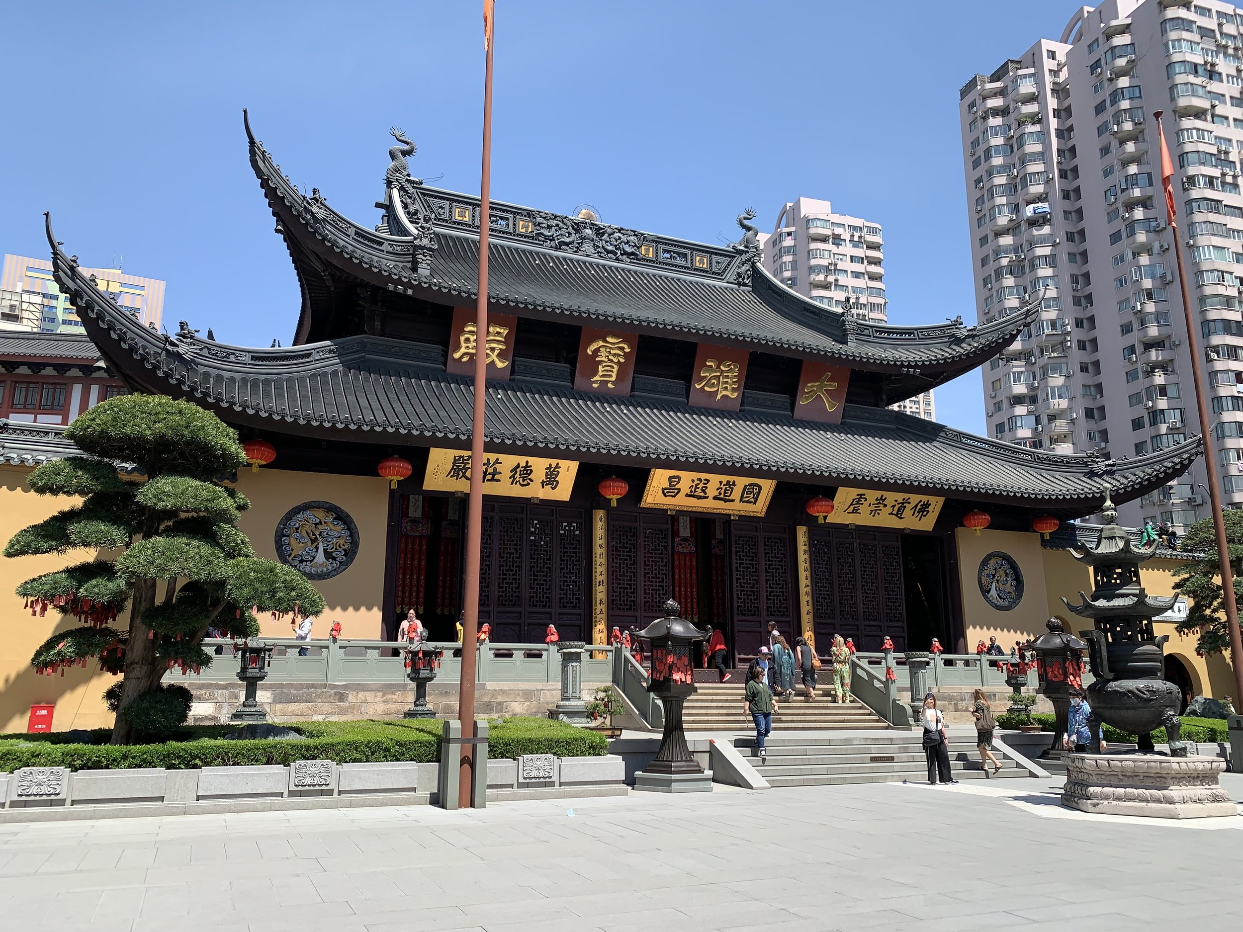 Traditional Chinese-style temple with ornate roof and golden Chinese characters on red banners, surrounded by modern high-rise buildings and people visiting on a sunny day.
