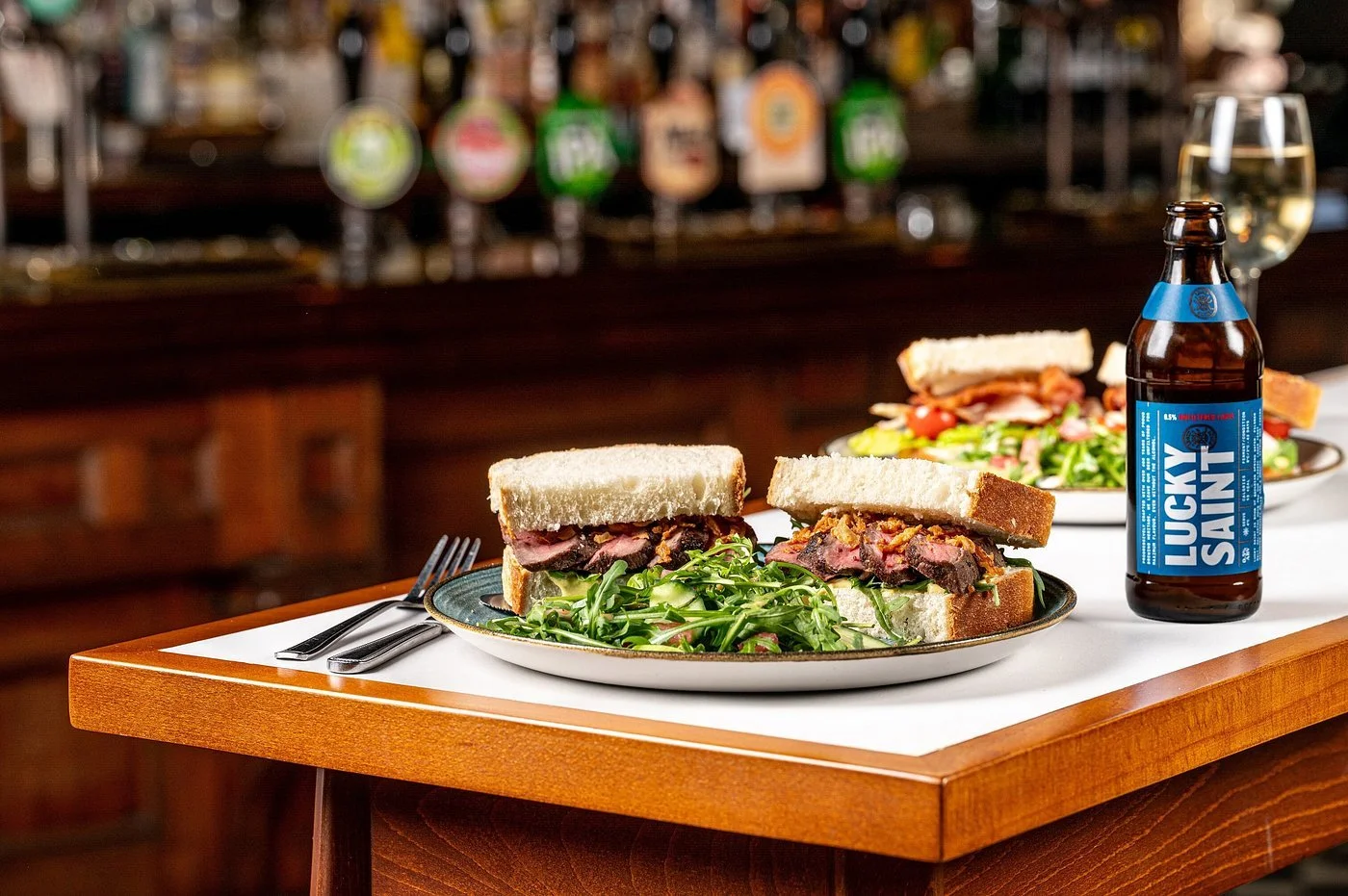 Sandwiches with sliced beef and greens on a plate, beer with a blue label, and a glass of white wine on a white tablecloth in a bar or restaurant setting.
