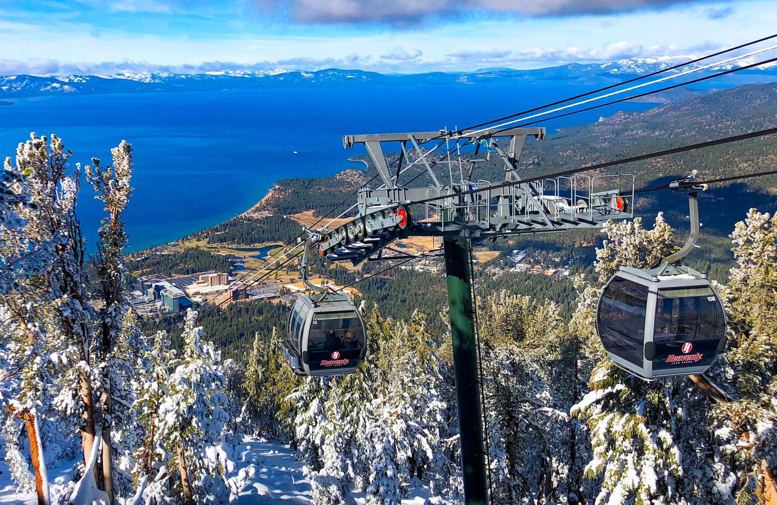 Cable car gondolas traveling over snow-covered trees on a mountain with a large lake and distant mountains in the background.